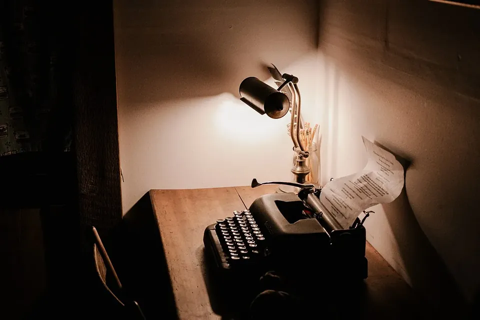A manual typewriter with a sheet of paper on a wooden desk, lit by a small desk lamp in a dark room