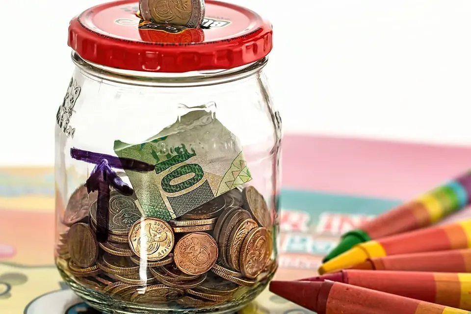 A glass jar with a red lid full of coins and a folded banknote, beside a pile of crayons