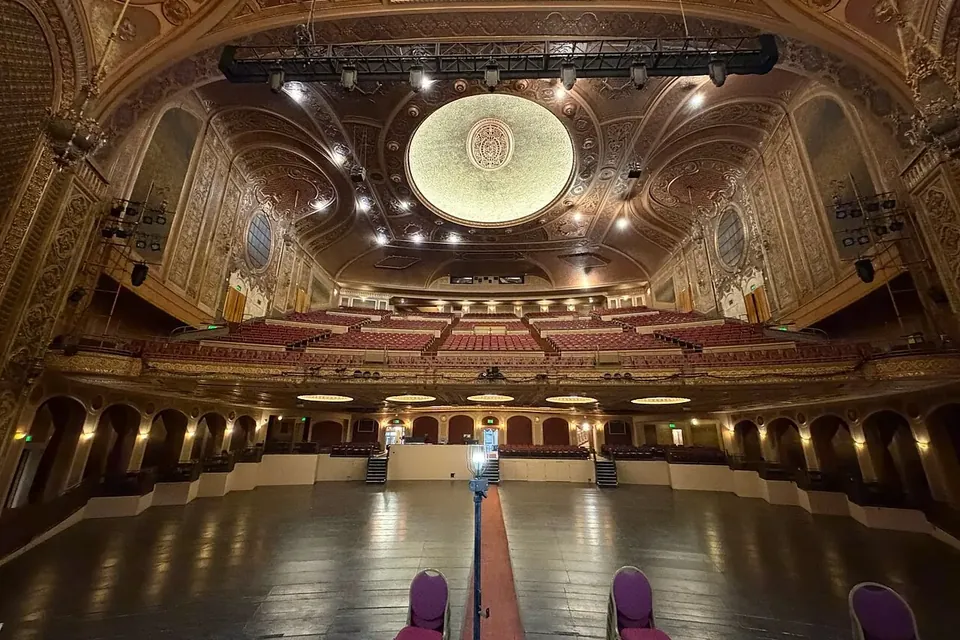 An empty, ornate theatre auditorium seen from the stage, with rows of red seats under a gilded domed ceiling