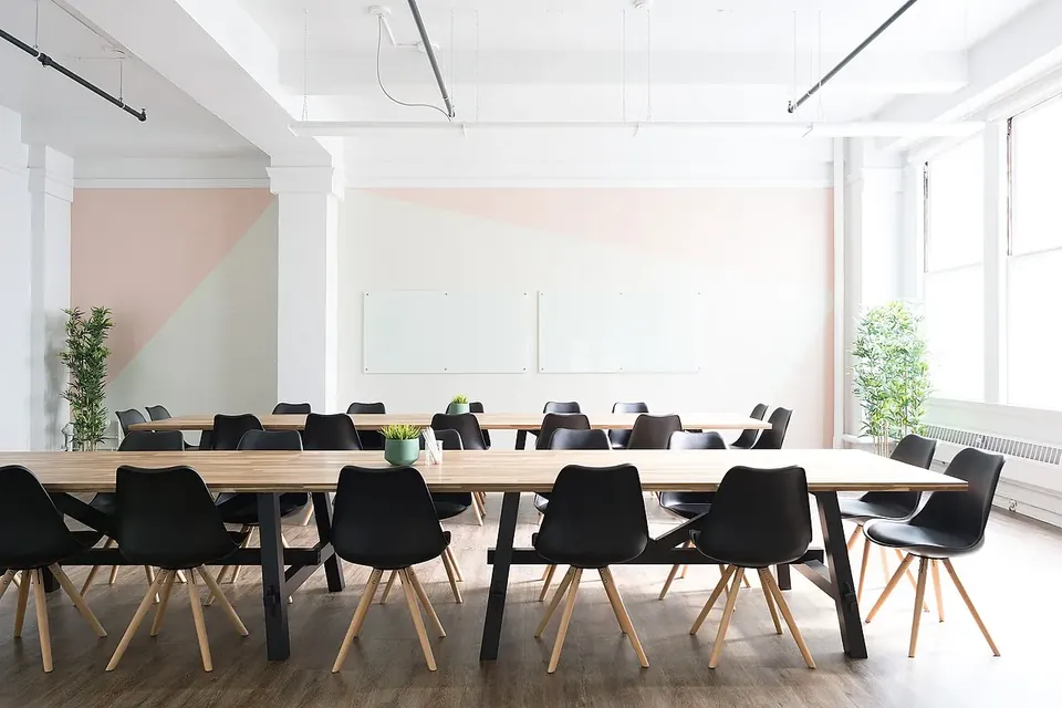 A bright, empty meeting room with long wooden tables and rows of black chairs