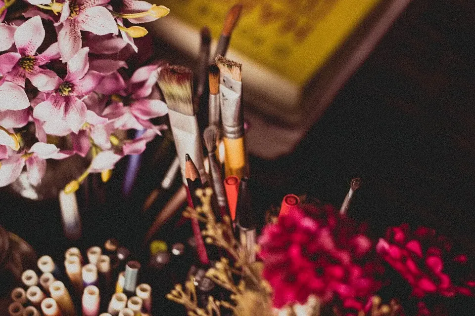 Paintbrushes, pencils and felt pens standing in pots on a cluttered desk, beside a vase of pink flowers