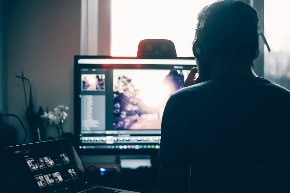 A person seen from behind wearing headphones, working at a desk with a large monitor and a laptop